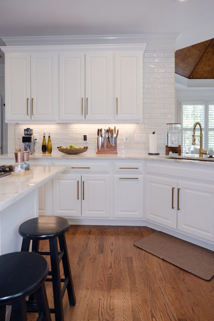 White raised-panel kitchen with subway tile, gold pulls, veined quartz counters, and black wood barstools.