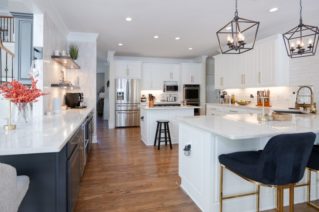 Open two-tone kitchen with white cabinets, a navy run, brushed-gold hardware, veined quartz counters, and lantern pendants.