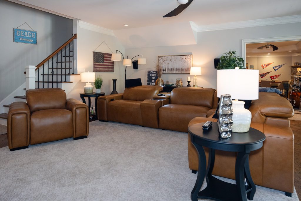 Basement media lounge with tan leather recliners, staircase, American-flag and beach decor, and warm lamplight.
