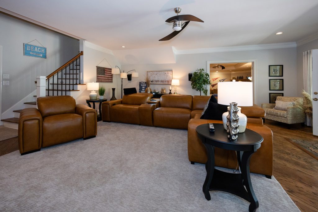 Finished basement with caramel leather recliners, area rug, table lamps, staircase, and a beach sign accent.