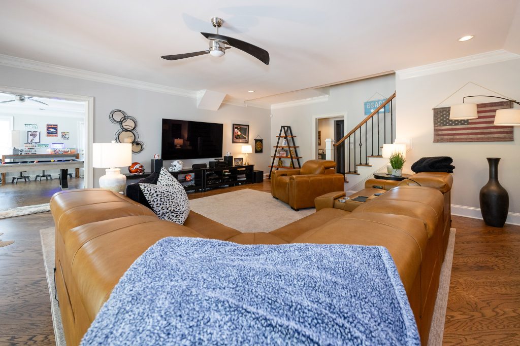 Basement lounge with tan leather sectional, mounted TV, ceiling fan, American-flag art, and a game room beyond.