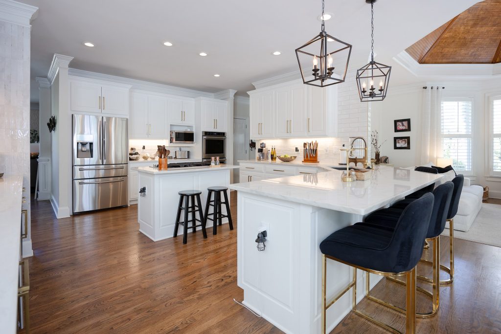 White kitchen with two quartz-topped islands, navy velvet barstools, lantern pendants, and stainless appliances.