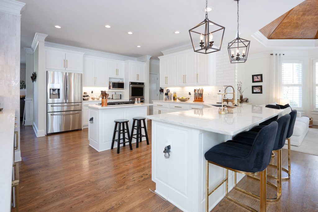 Two-island white kitchen with veined quartz, navy velvet barstools, gold legs, and iron lantern pendants.
