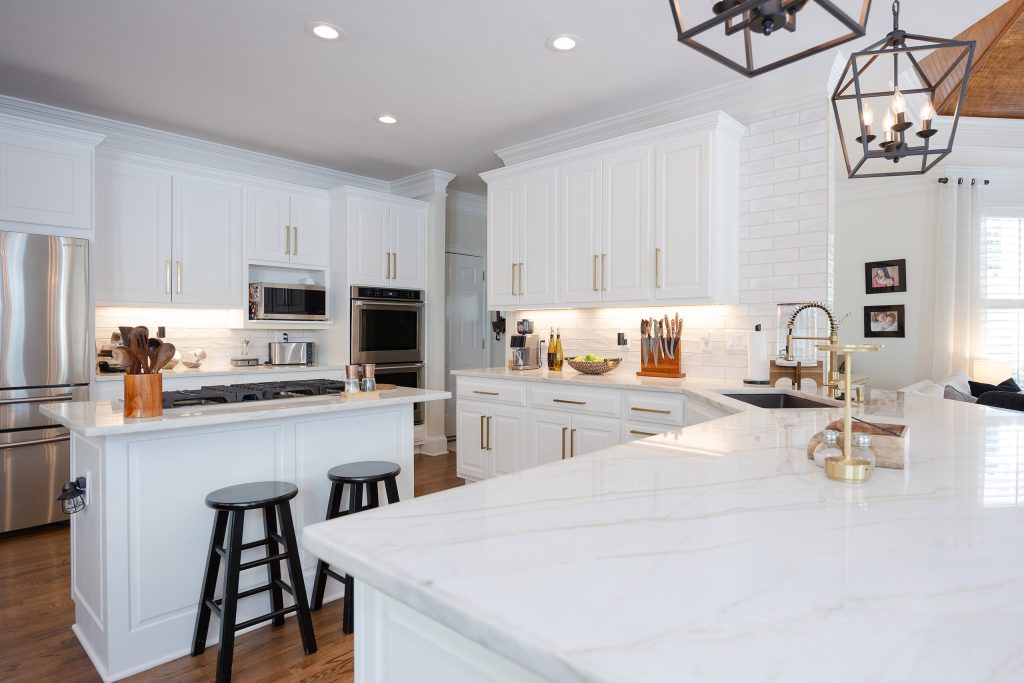 White kitchen with veined quartz waterfall counter, gold faucet, black stools, subway tile, and cage pendants.