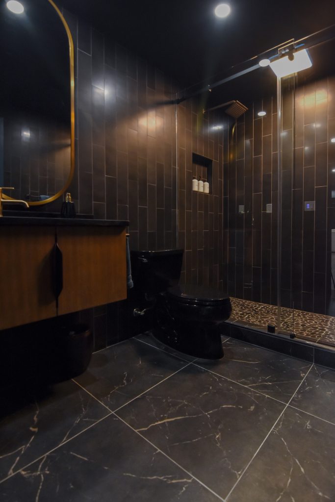 Lower-level bathroom with a floating wood vanity, gold mirror, and a walk-in shower with a pebble tile floor