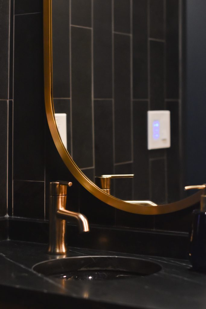 Round gold mirror and brushed gold faucet above a black undermount sink in a dark tiled bathroom