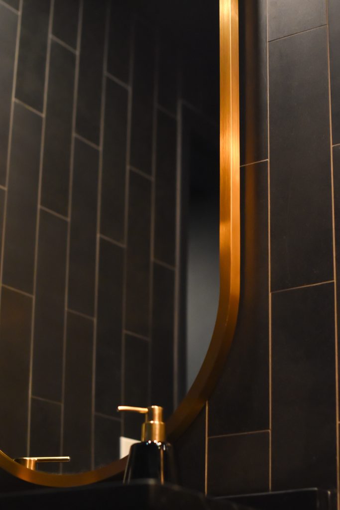 Brushed gold mirror frame and soap dispenser against vertical black wall tile