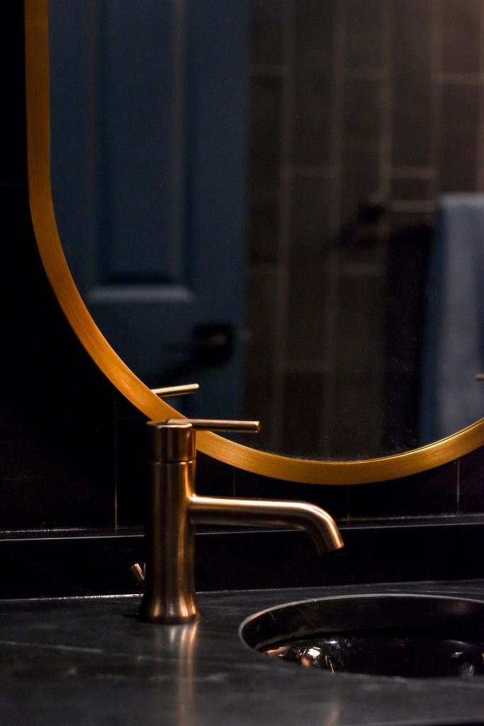 Brushed gold faucet and round mirror above a black undermount sink and dark stone vanity top