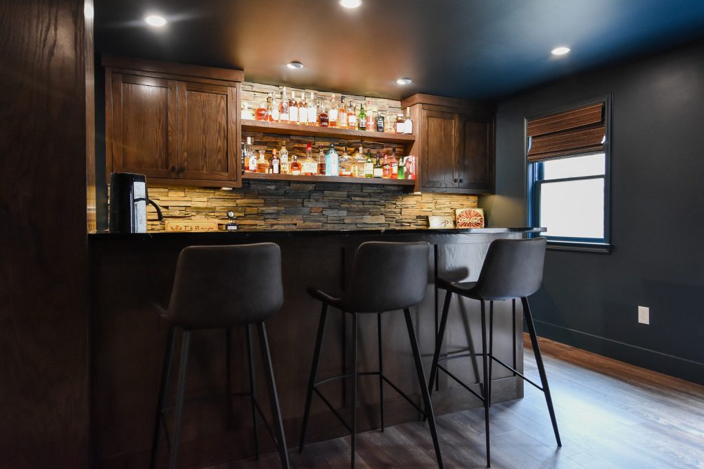 Custom basement wet bar in Cumming, GA with dark stained cabinetry, stacked stone backsplash, and leather bar stools