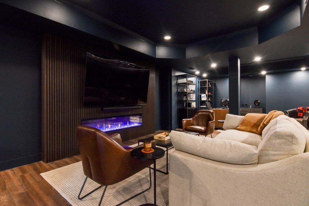 Finished basement living area with a fireplace media wall, leather chairs, and a home office beyond