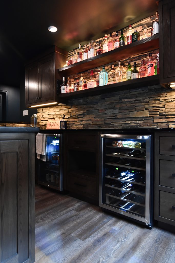 Backlit bottle shelving and stacked stone backsplash above a built-in wine refrigerator in a basement bar