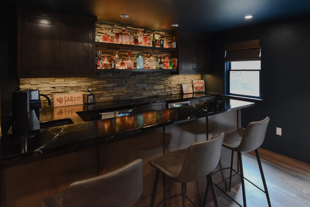 Wet bar counter and backlit shelving in a finished basement, with stacked stone and dark wood cabinetry