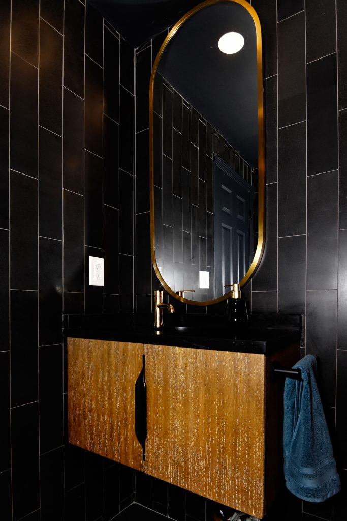Basement bathroom with vertical black wall tile, a gold oval mirror, and a wood-front floating vanity