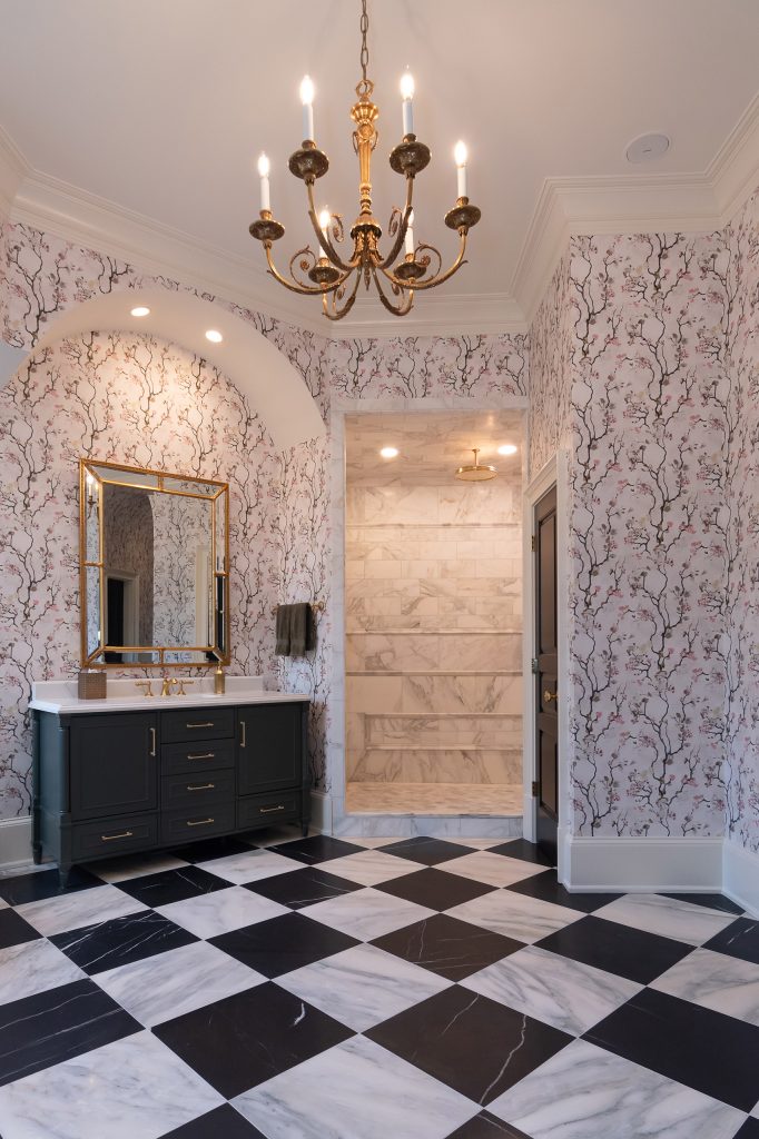 Charcoal vanity with gilded mirror beside a marble walk-in shower with gold rain head under a candelabra chandelier.