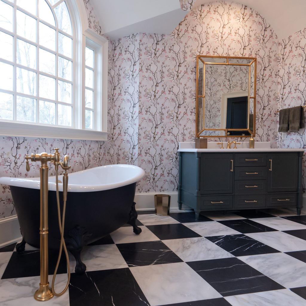 Black clawfoot tub under an arched window beside a charcoal vanity with gold mirror in a blossom-papered bath.