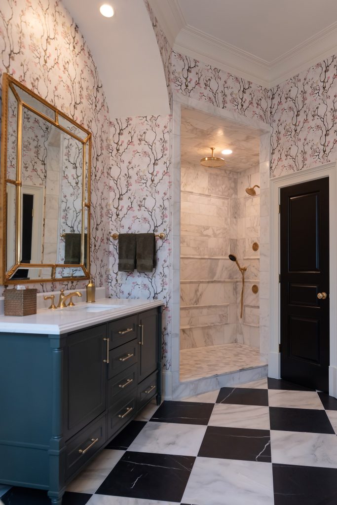 Charcoal vanity with gold mirror beside a marble walk-in shower and a black paneled door on checkerboard marble.