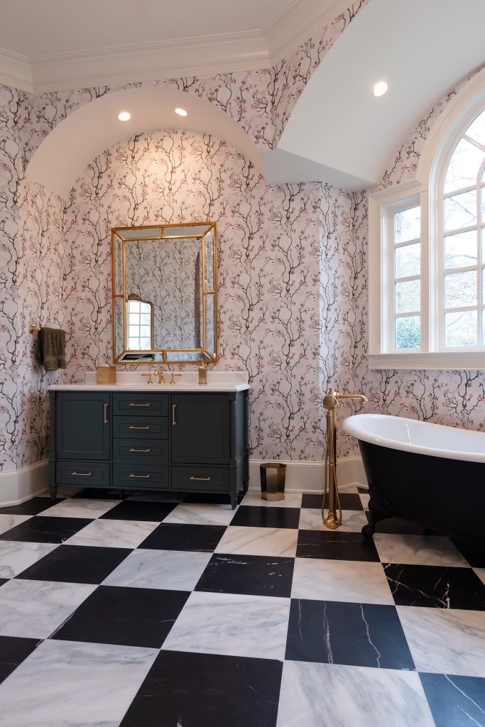 Charcoal vanity and black clawfoot tub in a vaulted blossom-papered bath with arched window and gold mirror.