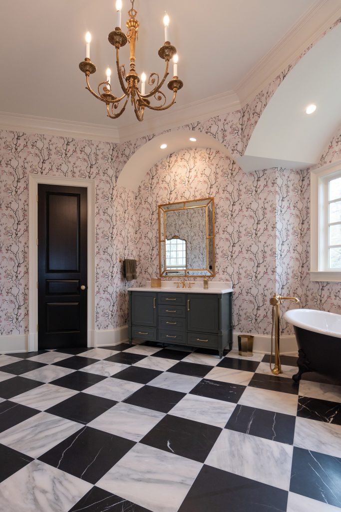 Charcoal vanity with gold mirror in an arched niche, gilded chandelier, and black clawfoot tub on checkerboard marble.