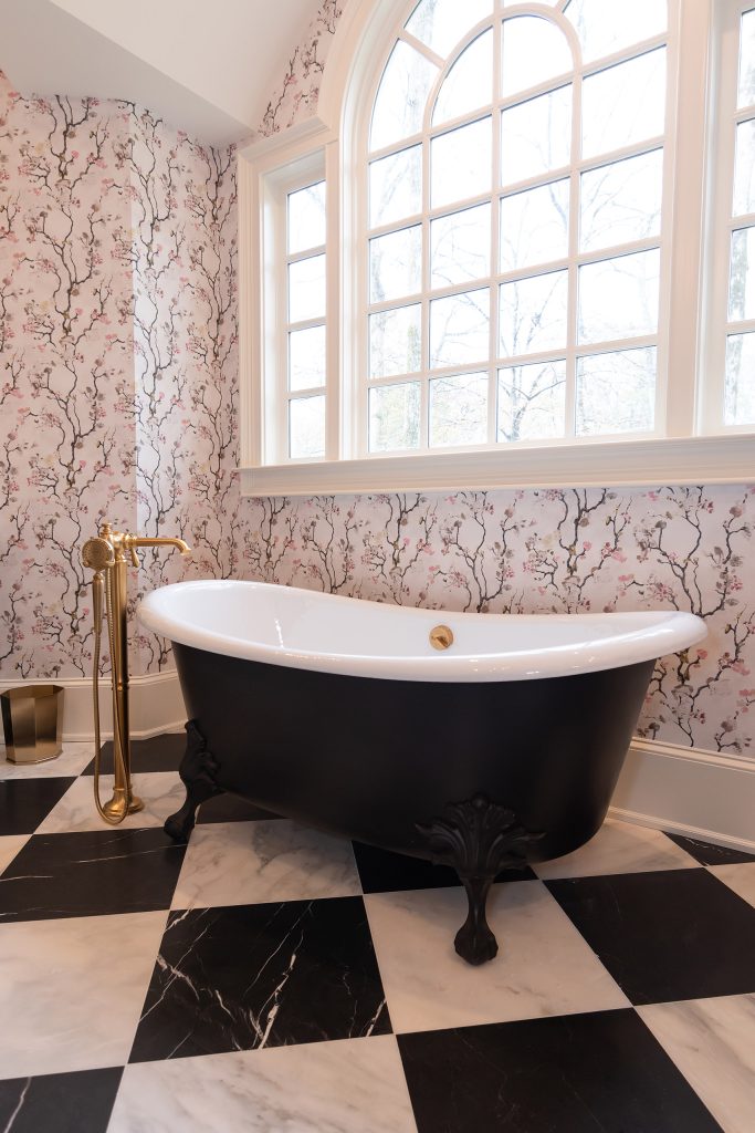 Black slipper clawfoot tub with gold filler beneath a large arched divided-light window, on marble checkerboard.
