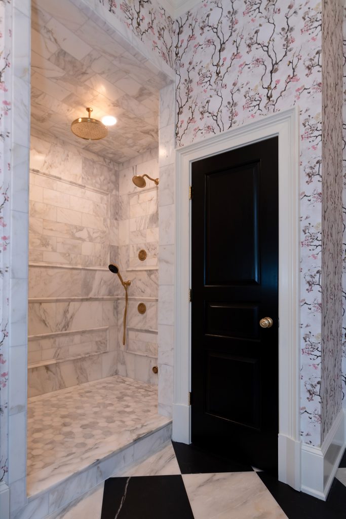 Marble walk-in shower with gold rain head and hex floor beside a glossy black paneled door with glass knob.