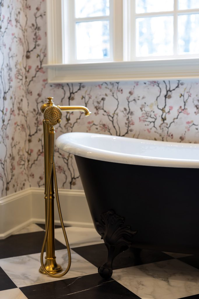 Black cast-iron slipper clawfoot tub with brushed-gold floor filler under a window, on checkerboard marble.