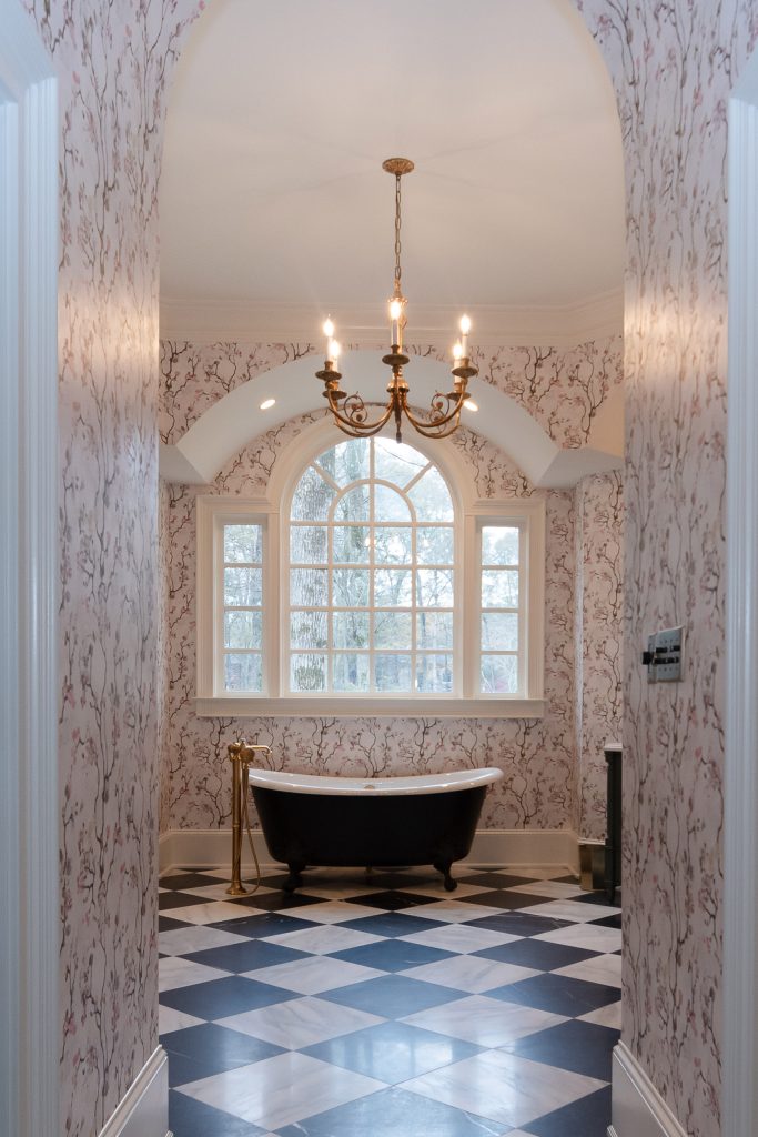Doorway view into a vaulted bath with black clawfoot tub below an arched window and gold chandelier.