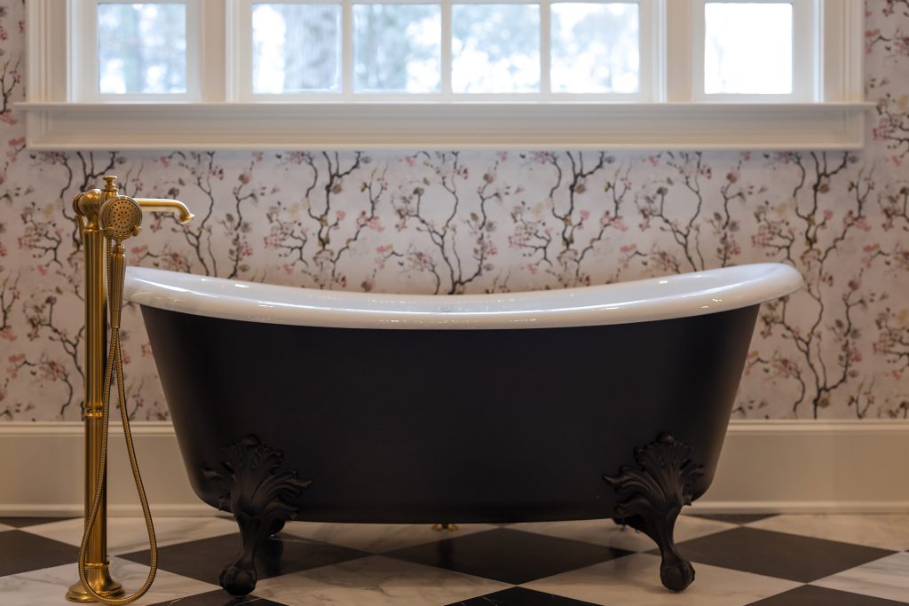Black cast-iron slipper clawfoot tub with brushed-gold filler beneath a window, on black-and-white checkerboard marble.