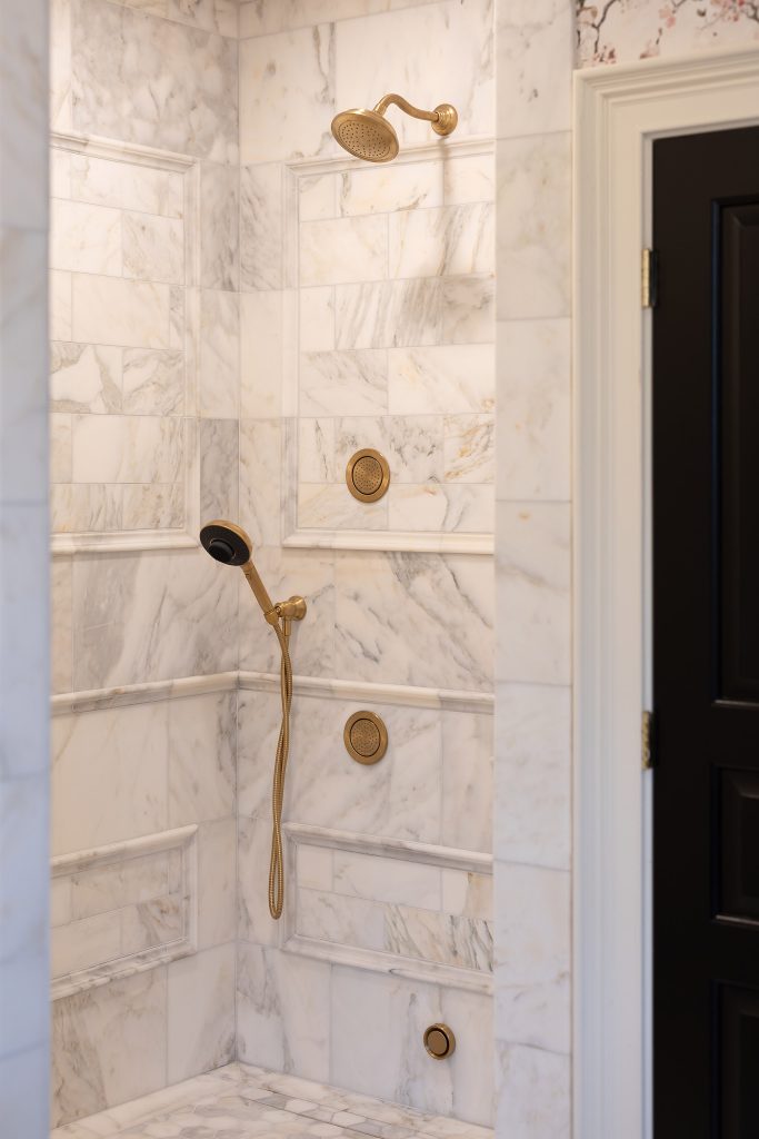 Marble shower wall with brushed-gold showerhead, handheld wand, and round body jets framed by carved tile trim.