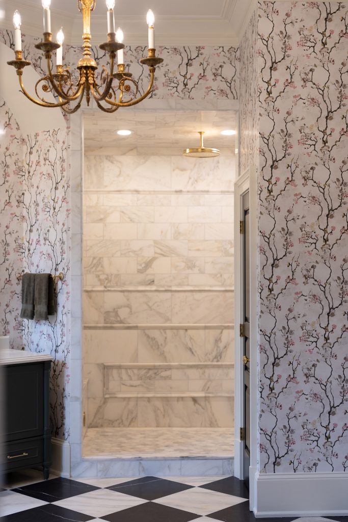 Marble walk-in shower framed by blossom-papered walls and a gilded candle chandelier above.