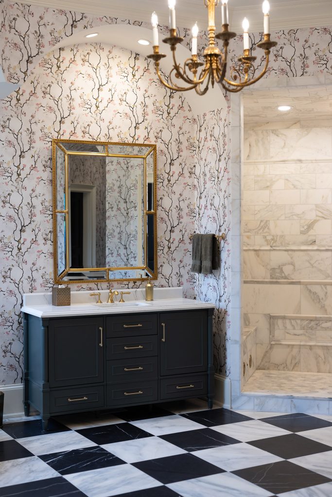 Charcoal vanity with gold mirror under a candelabra chandelier, next to a marble shower with bench.