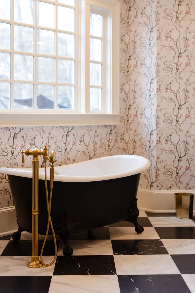 Charcoal furniture-style vanity with white quartz top, gold faucet, and a large gilded beveled mirror on blossom wallpaper.