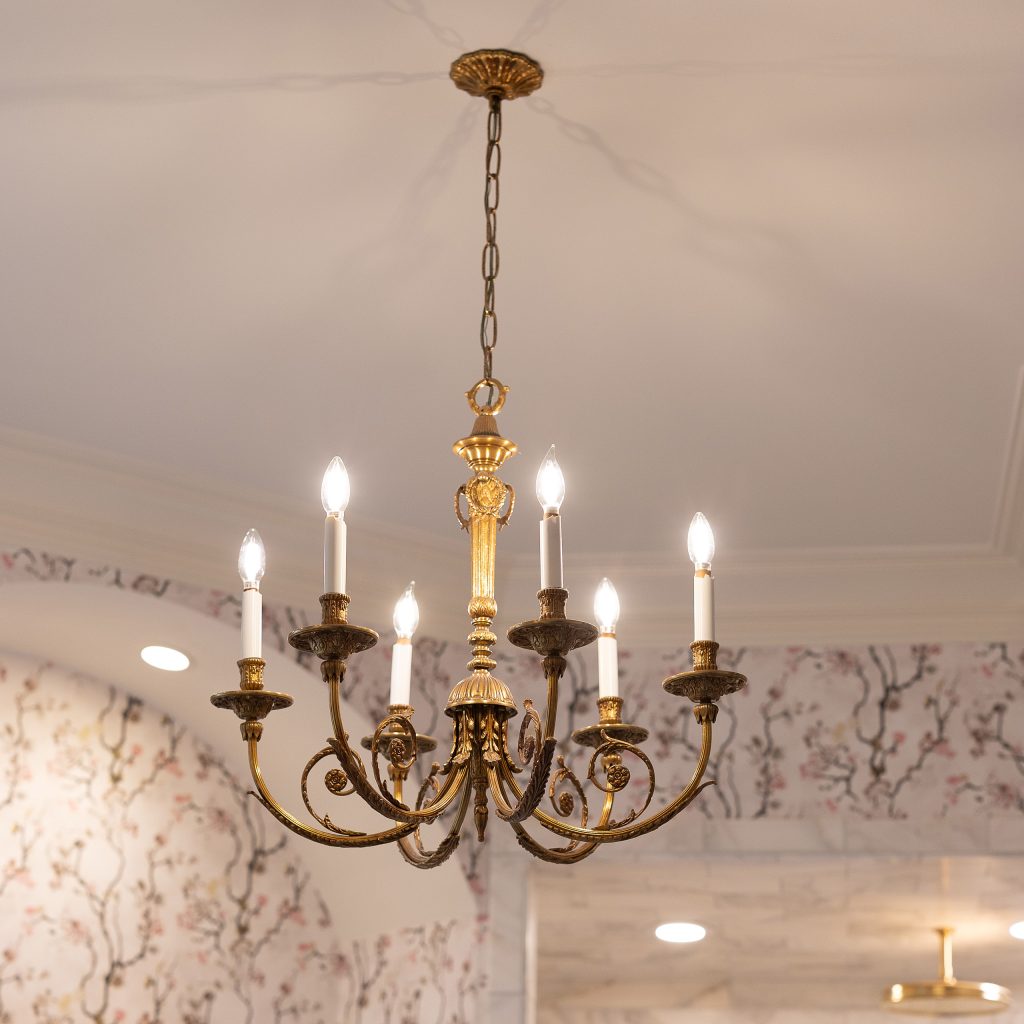 Antique gilded six-arm candelabra chandelier hanging from a coffered ceiling above blossom-print walls.