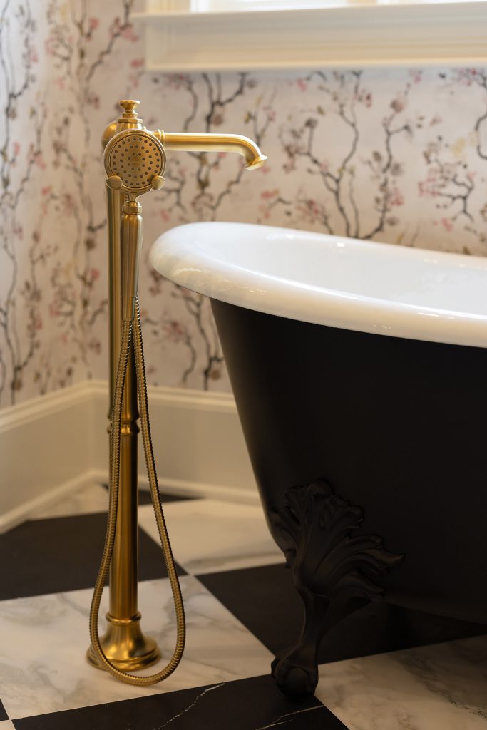 Brushed-gold floor-mounted tub filler with handshower beside the ornate cast-iron foot of a black slipper tub.