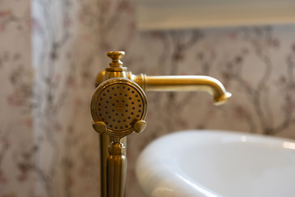Close-up of a brushed-gold floor-mounted tub filler and handshower against blossom wallpaper.