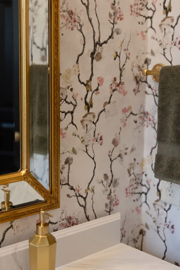 Corner of a gilded beveled mirror and gold soap pump against watercolor cherry-blossom wallpaper.