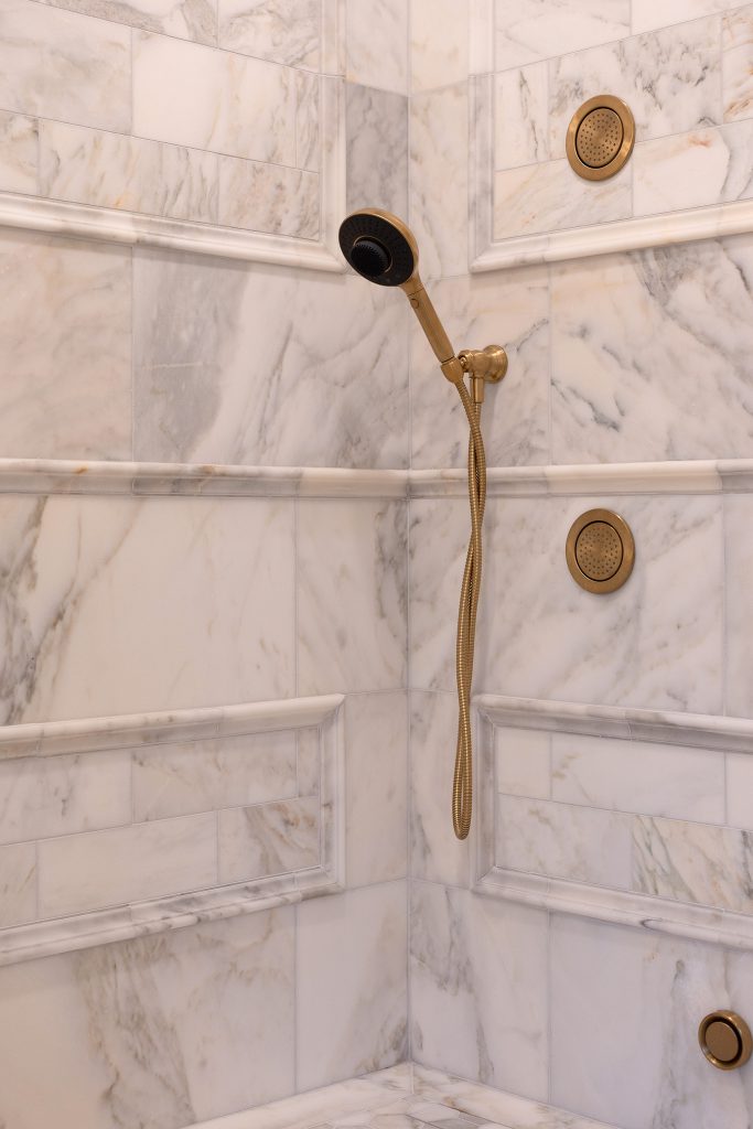 Close-up of shower corner with brushed-gold handheld wand, coiled hose, and two round body sprays on veined marble.