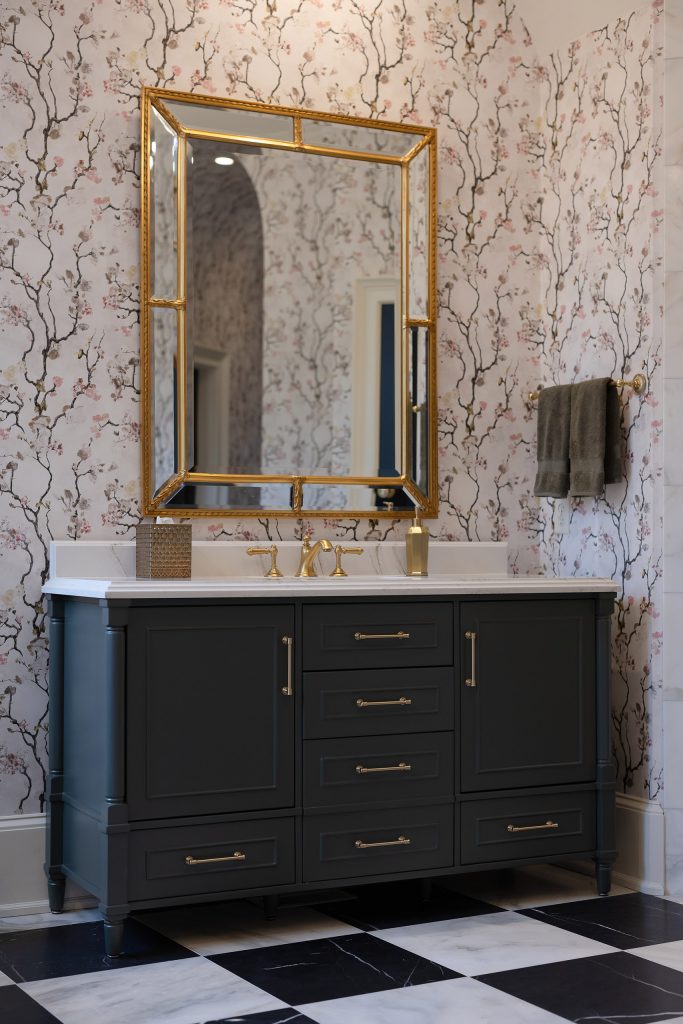 Vaulted blossom-papered bath with black paneled door, charcoal vanity, and gold chandelier over checkerboard marble.
