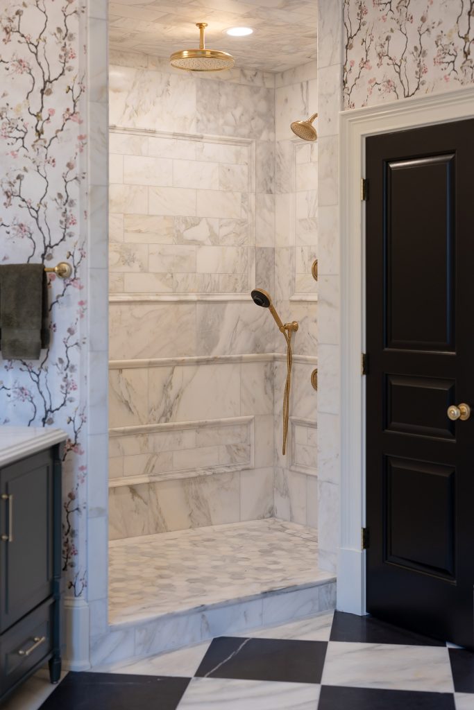 Marble-tiled walk-in shower with brushed-gold rain head, handheld, and body sprays beside a black paneled door.
