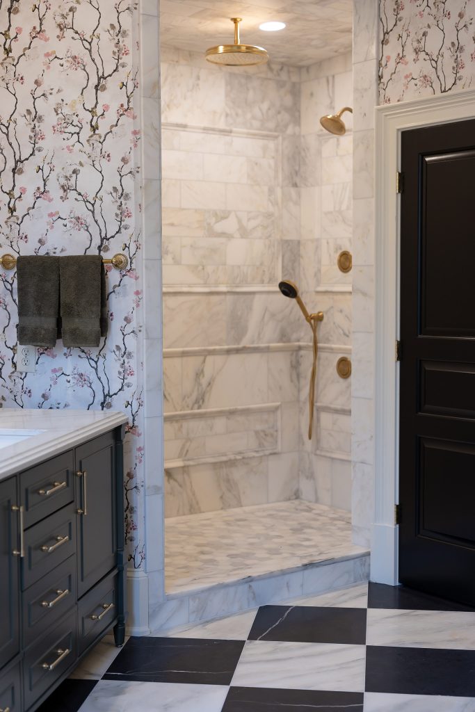 Charcoal vanity beside a marble walk-in shower with gold rain head, framed by blossom wallpaper and a black door.