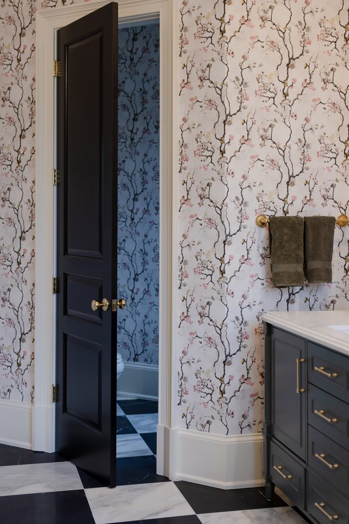 Open glossy black paneled door with glass knob beside blossom wallpaper, olive towels, and a charcoal vanity.