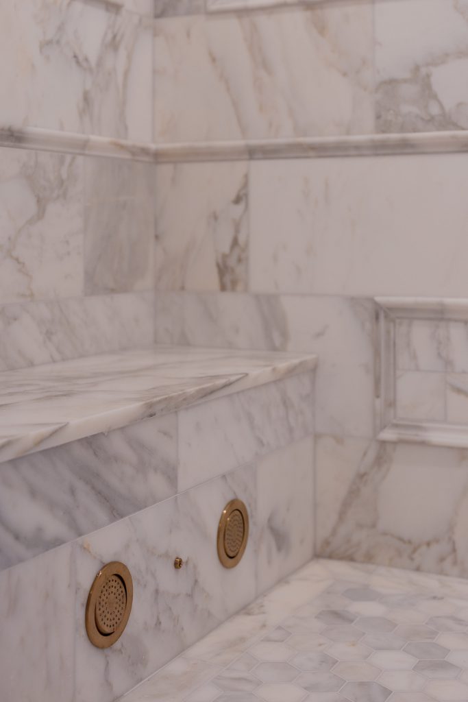 Close-up of a marble shower bench and framed niche with brushed-gold body-spray jets and a hex marble floor.