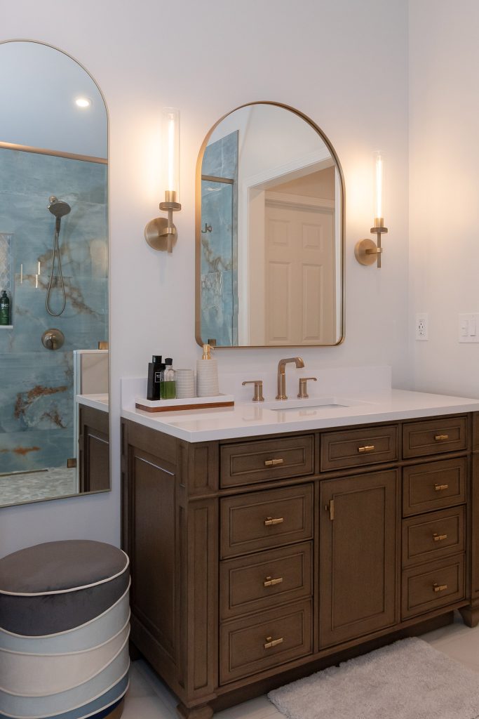 Dark-wood vanity with white quartz under a tall arched mirror that reflects the blue onyx shower, with a striped ottoman.