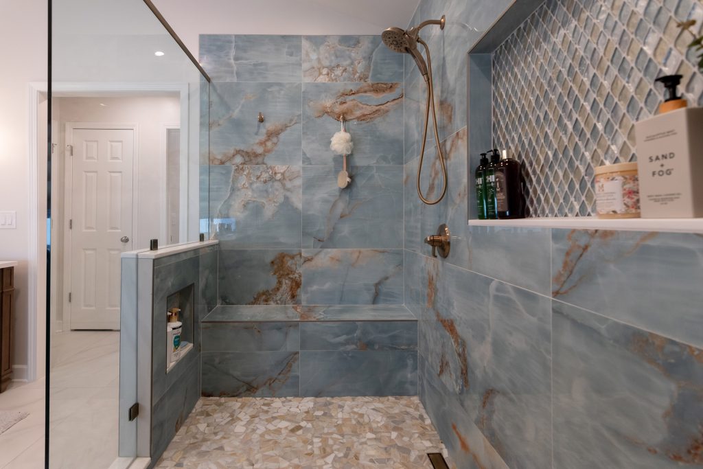 Blue onyx shower with amber veining, a diamond mosaic niche, a bench, and pebble floor, with a bathroom door beyond the glass.