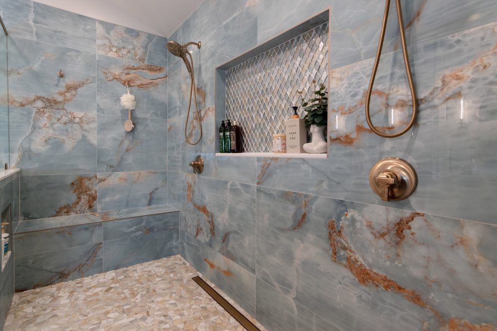 Close view of a blue onyx shower with rust-veined stone, a diamond mosaic niche, a tiled bench, and a river-pebble floor.