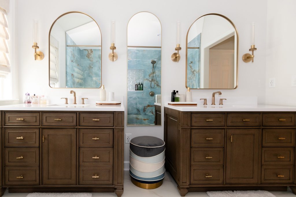Two dark-wood vanities facing off with arched brass mirrors, a striped blue ottoman between them, and the onyx shower reflected.