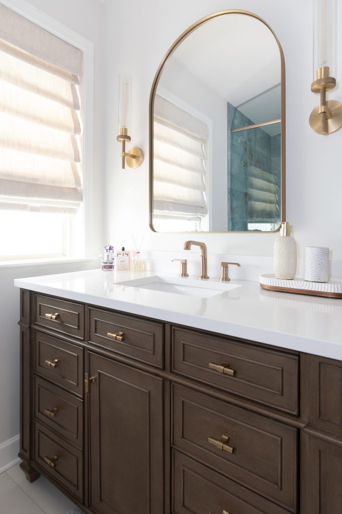 Single dark-wood vanity with white quartz and brushed-gold faucet beneath an arched mirror, next to a shaded window.