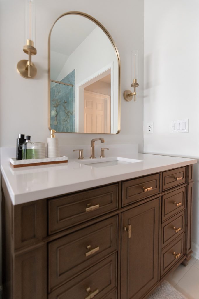Single dark-wood vanity with white quartz, arched brass mirror, and a tall side mirror reflecting the blue onyx shower.