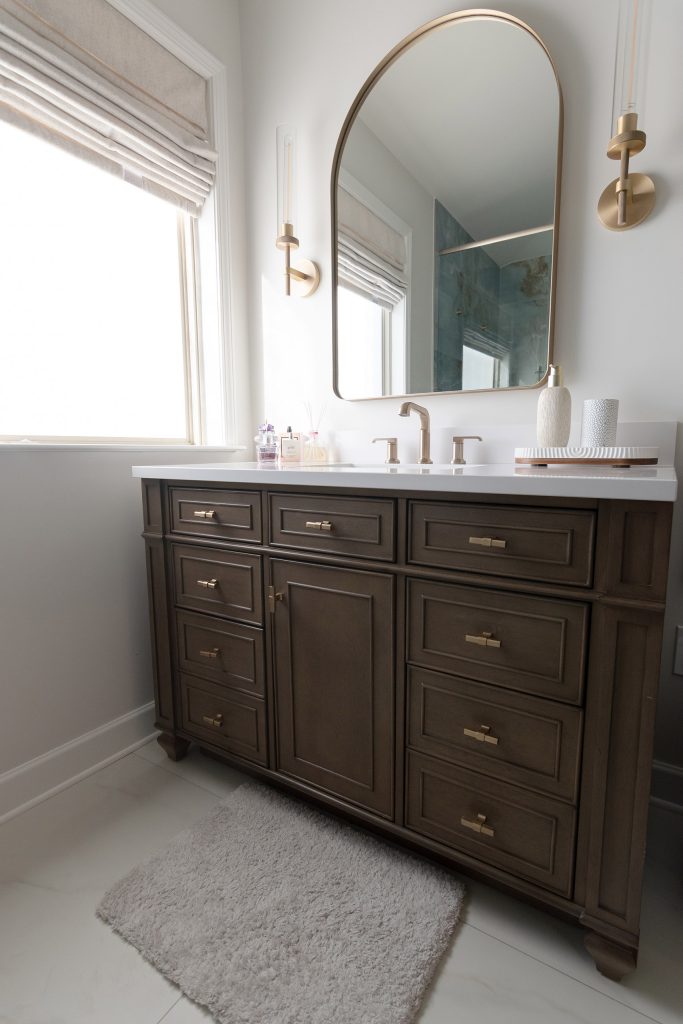 Single dark-wood vanity with a white quartz top, brushed-gold faucet, an arched brass mirror, and a shaded window.