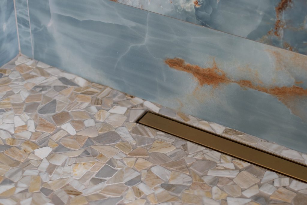 Detail of a pebble mosaic shower floor meeting blue onyx stone walls, with a slim brushed-gold linear drain between them.
