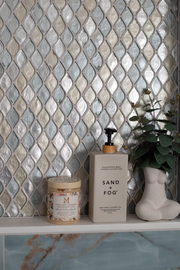 Shower niche detail with an iridescent teardrop glass mosaic back, a Sand + Fog bottle, and a eucalyptus sprig over blue onyx.